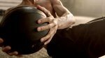 Muscular fit man performing medicine ball training drills inside a gym while topless