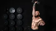 Shirtless man performing a full-body dumbbell workout in a packed gym