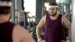 overweight man looking into the mirror in the gym trying to fix his bad habits